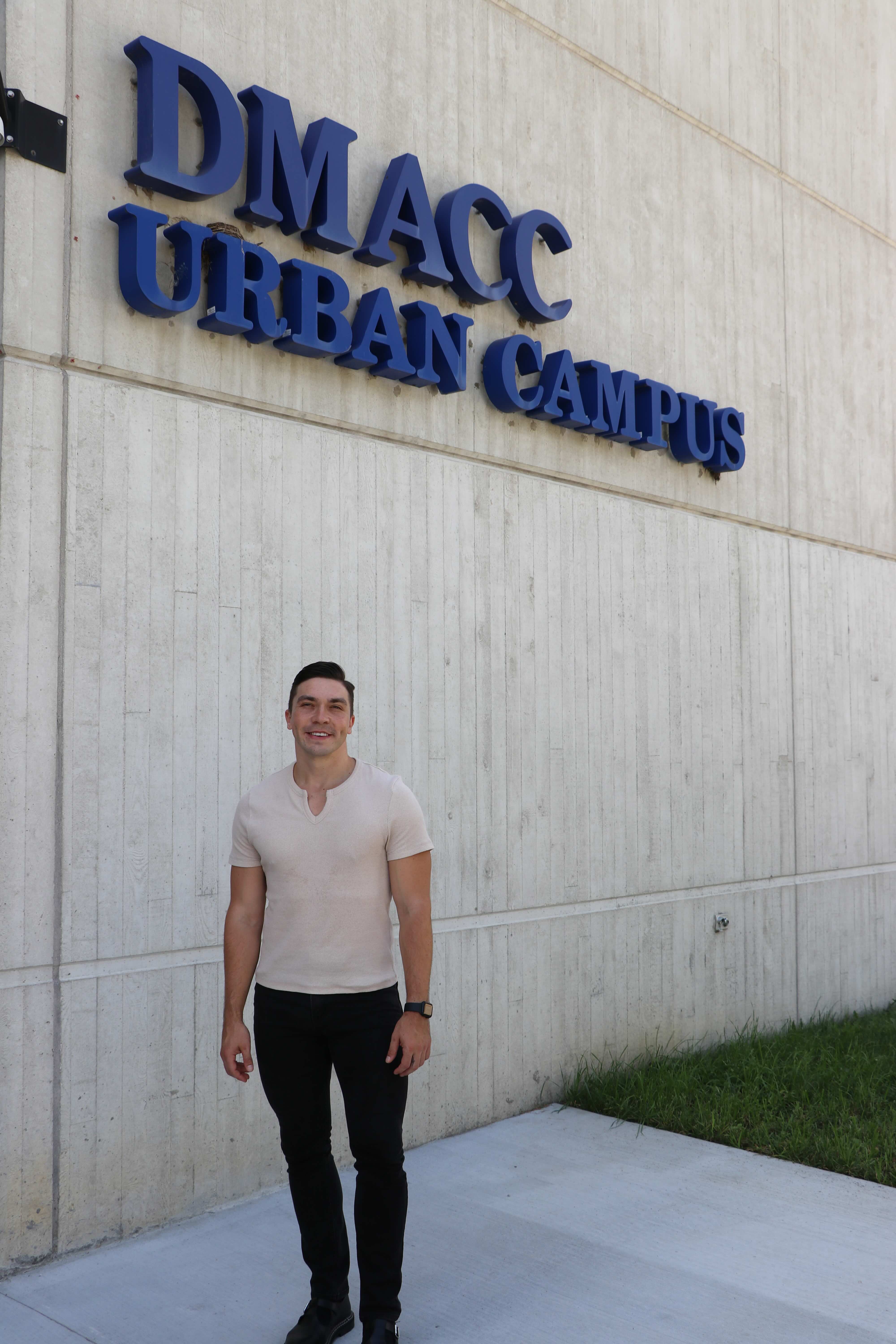 Man in white shirt and jeans stands in front of DMACC Urban Campus building 1 under a sign.