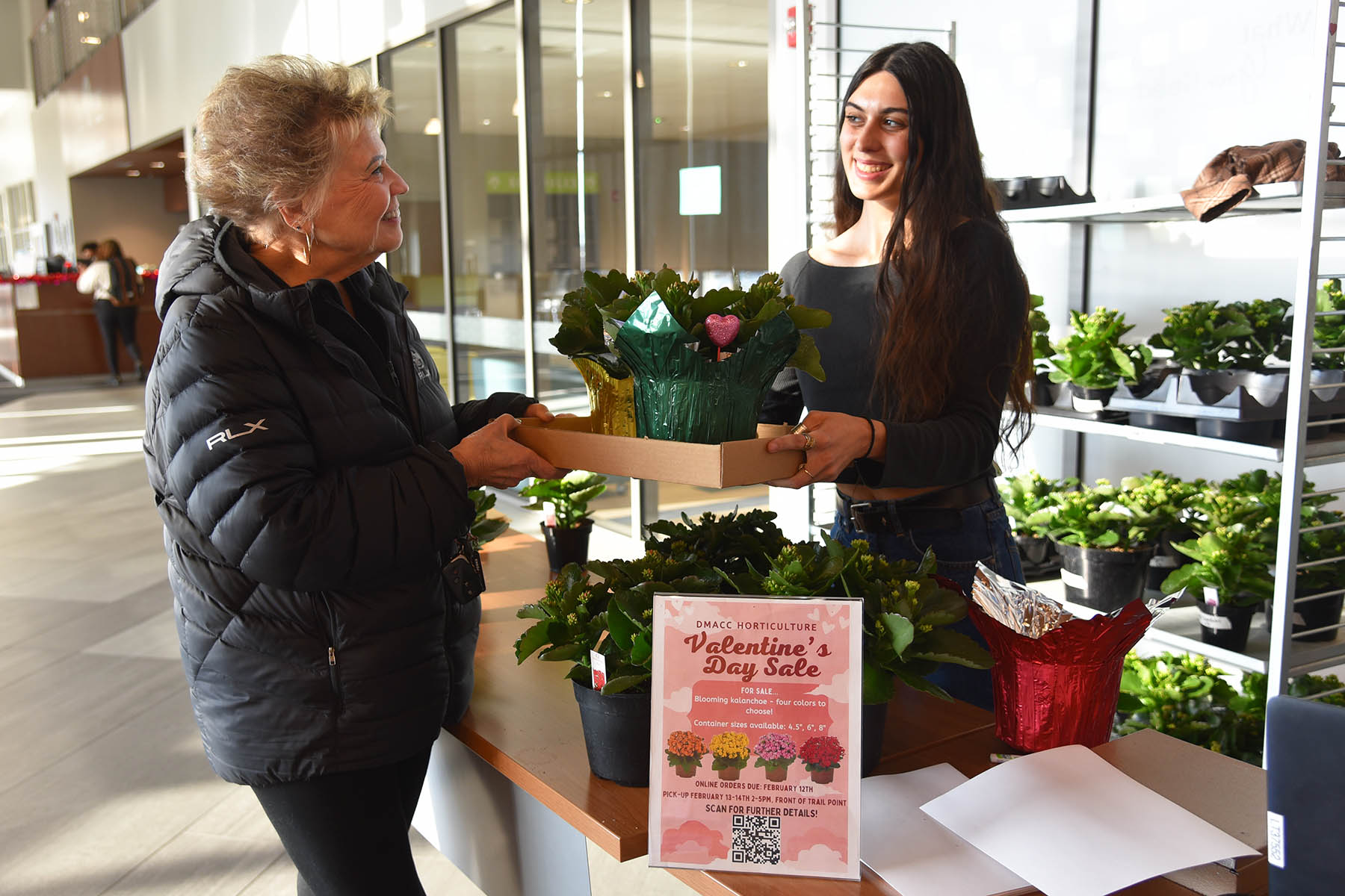 DMACC Horticult​​ure​ student Danii​ Brady (right) of Indianola and Nancy Nicodemus (left) of Urbandale share a smile as Brady hands Nicodemus her purchase during the first day of the DMACC Horticulture Club's Valentine's Day Sale on Tues., Feb. 13, at the College's Ankeny Campus.