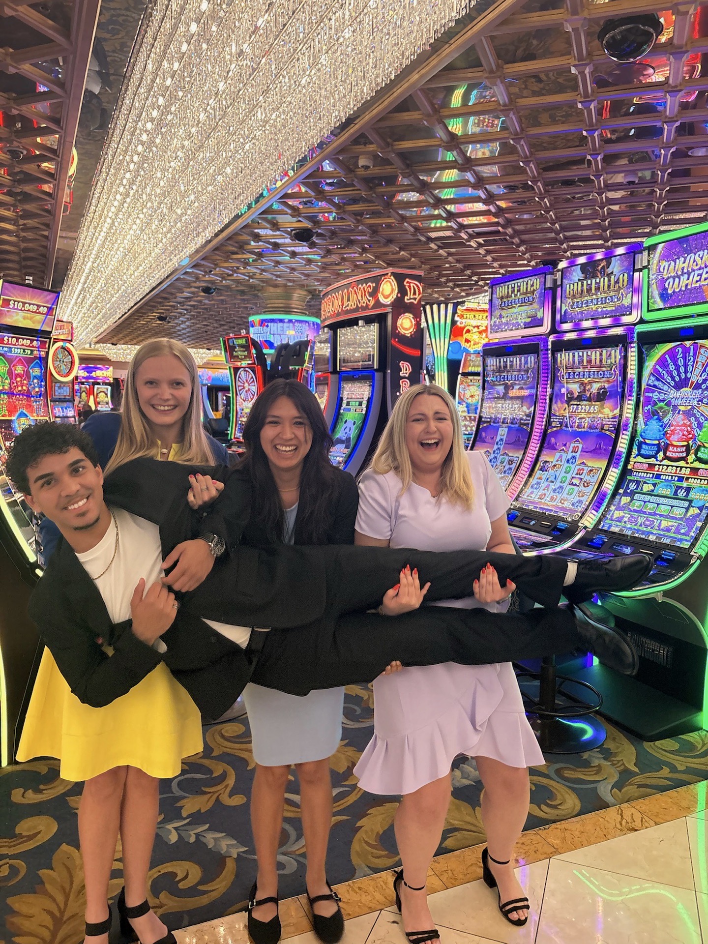 Three people carry a person horizontally posing for a photo in front of bright lights and slot machines