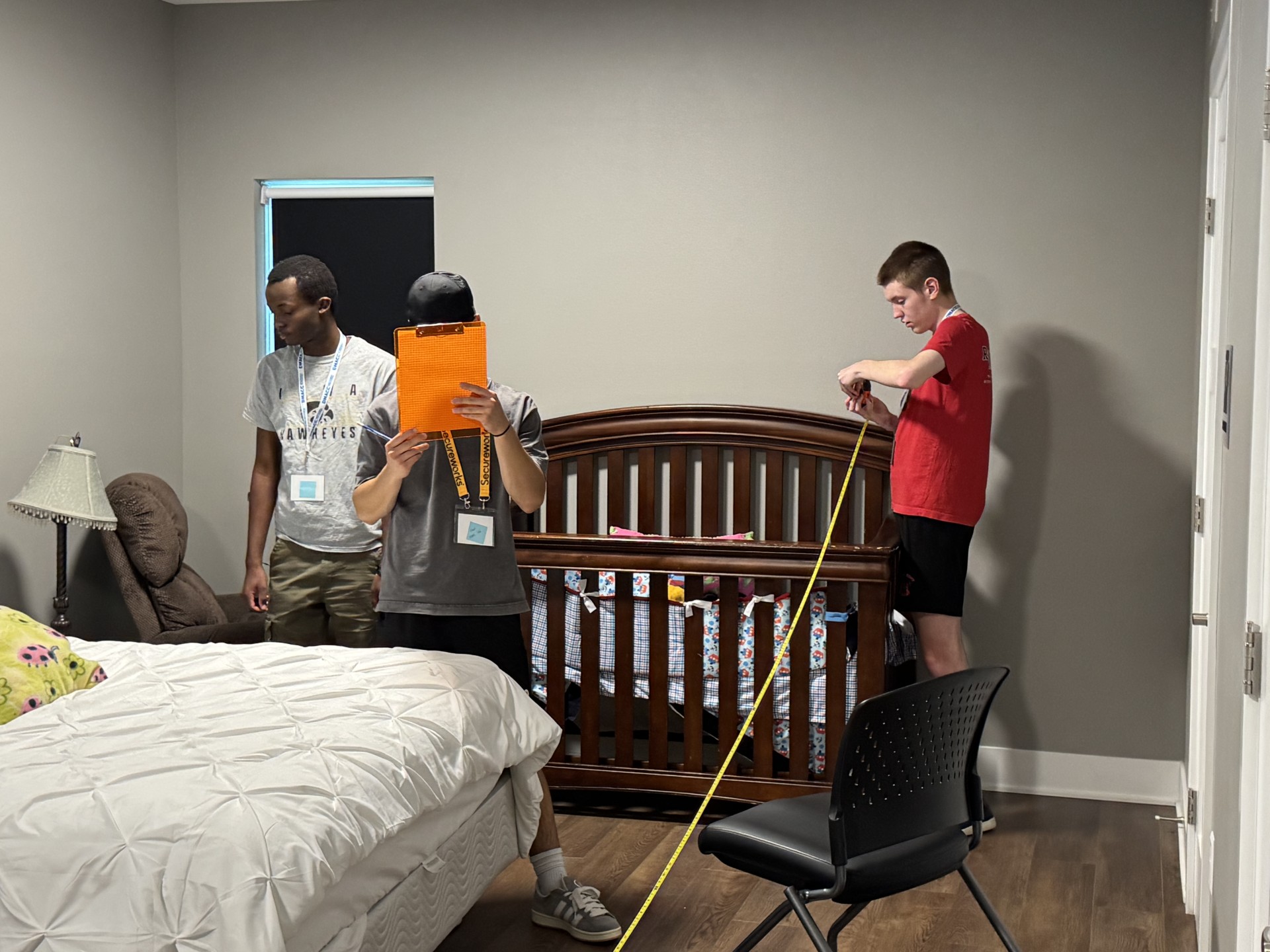 Three people stand in a bedroom with a bed and crib, holding measuring tape and a clipboard.