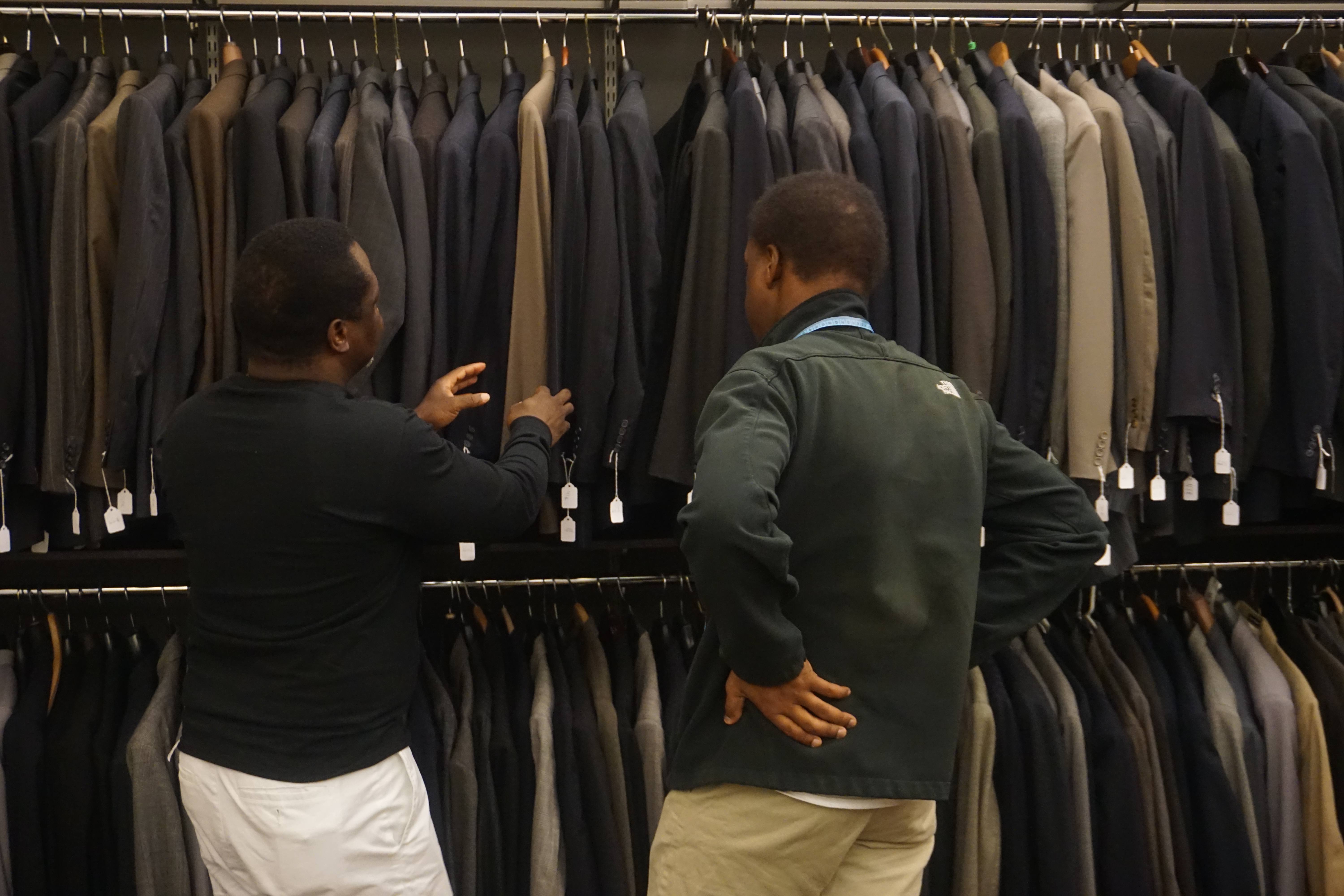 Two men face a rack of suits. 