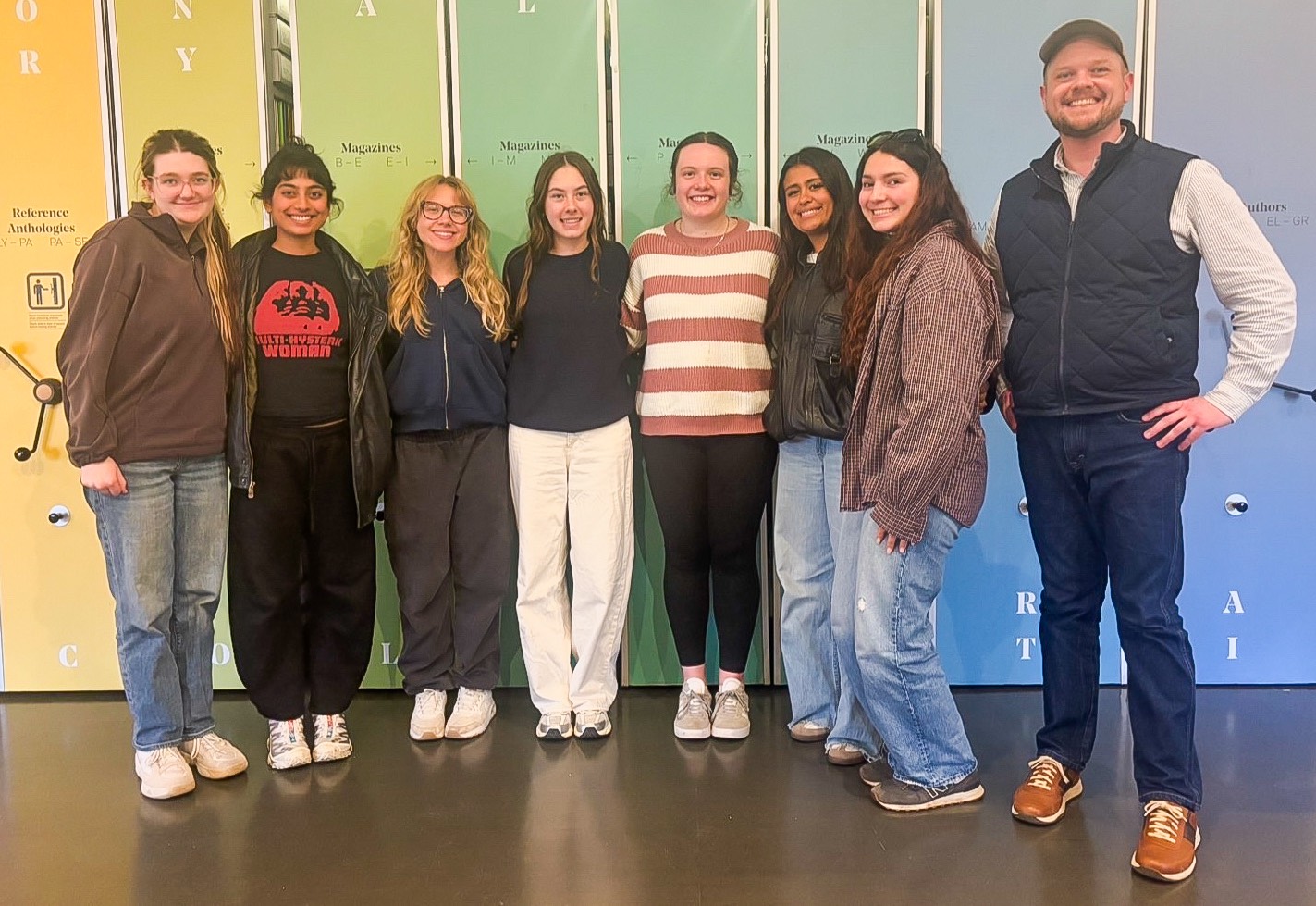 A group of eight casually dressed people stand next to each other for a photo against a colorful rainbow spectrum painted backdrop of book shelves.