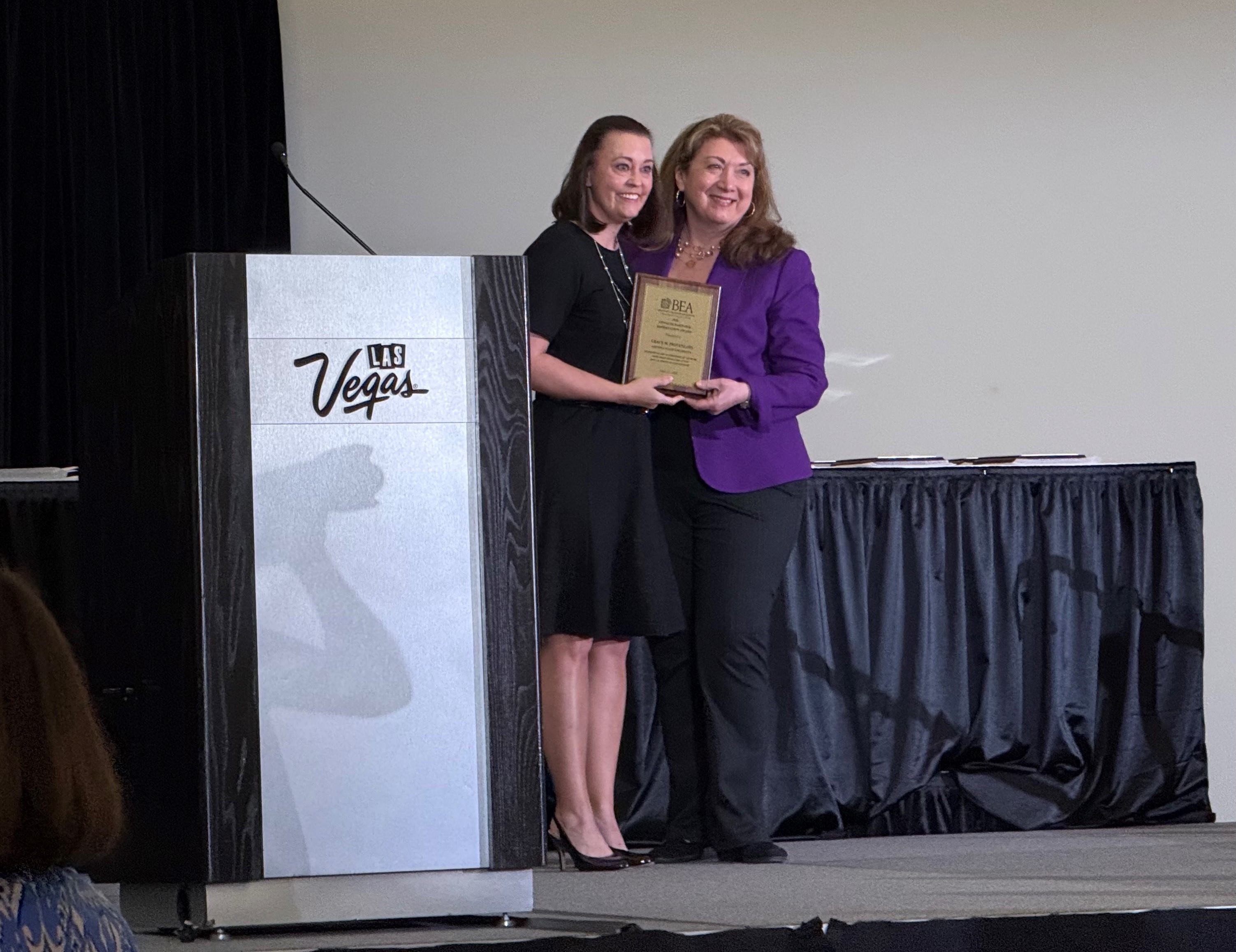 Two women in professional attire stand side by side holding a plaque next to a podium with Las Vegas as decal text on the front
