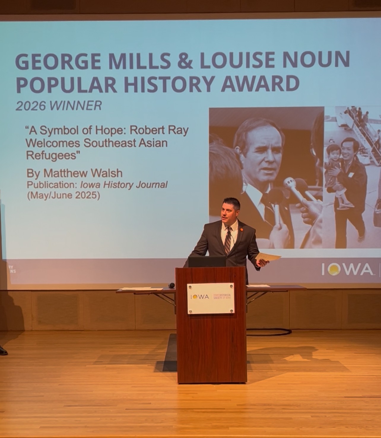 Man in suit stand at podium on stage with projected display of his award at the Iowa Historical Society Ceremony